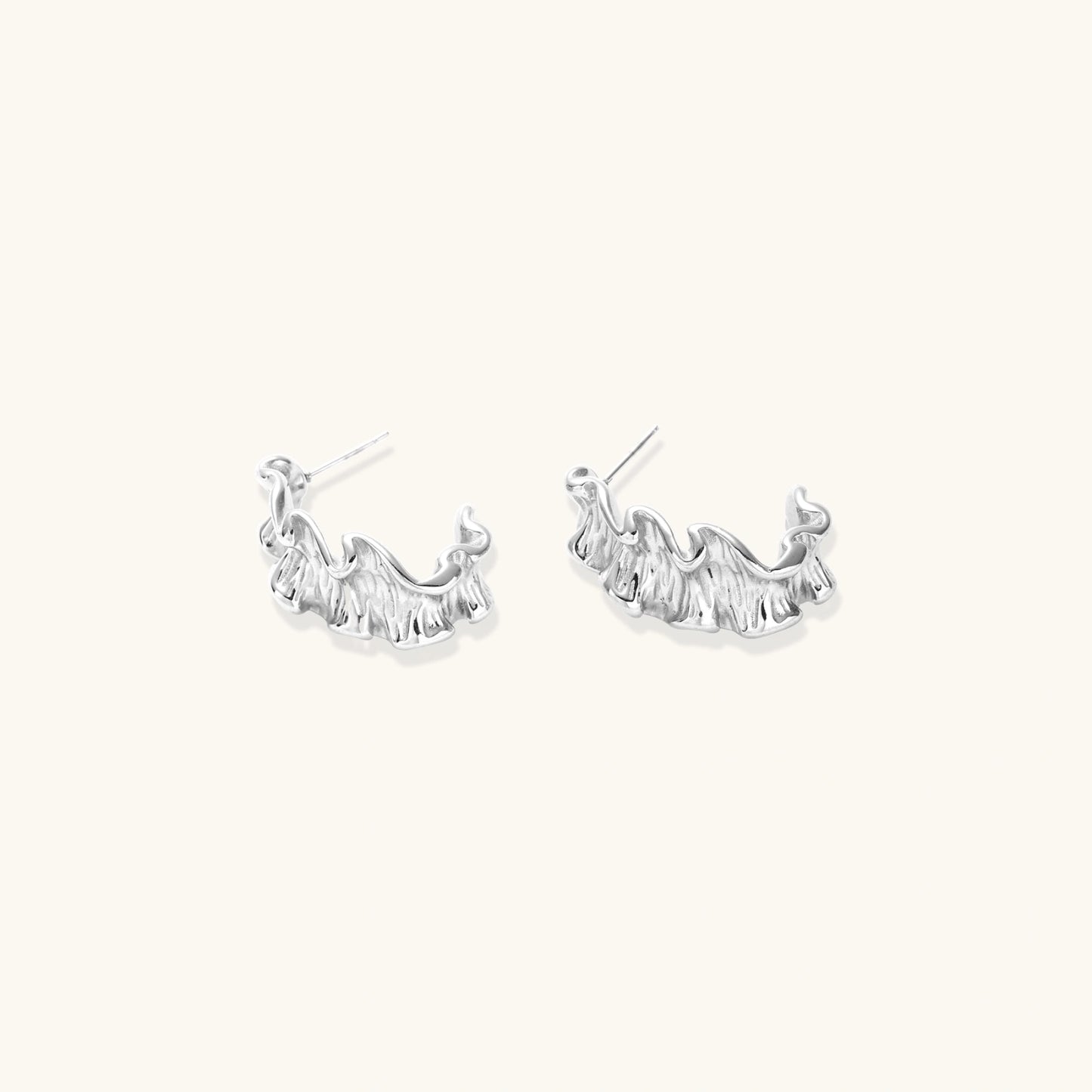 Textured white gold-plated hoop earrings with flowing wave pattern against cream background, showing dimensional rippled surface.